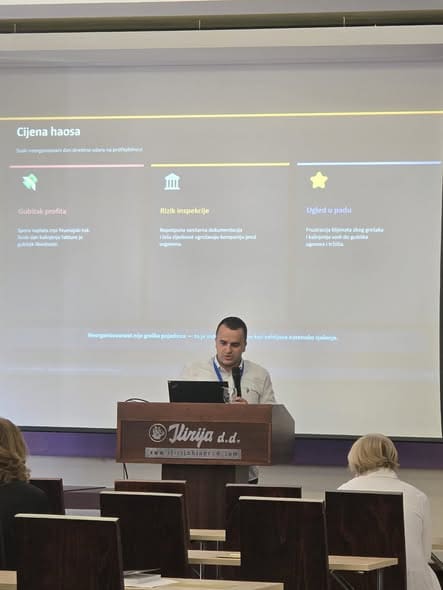 Muškarac za govornicom drži mikrofon tokom prezentacije, dok je iza njega prikazan slajd „Cijena haosa“.